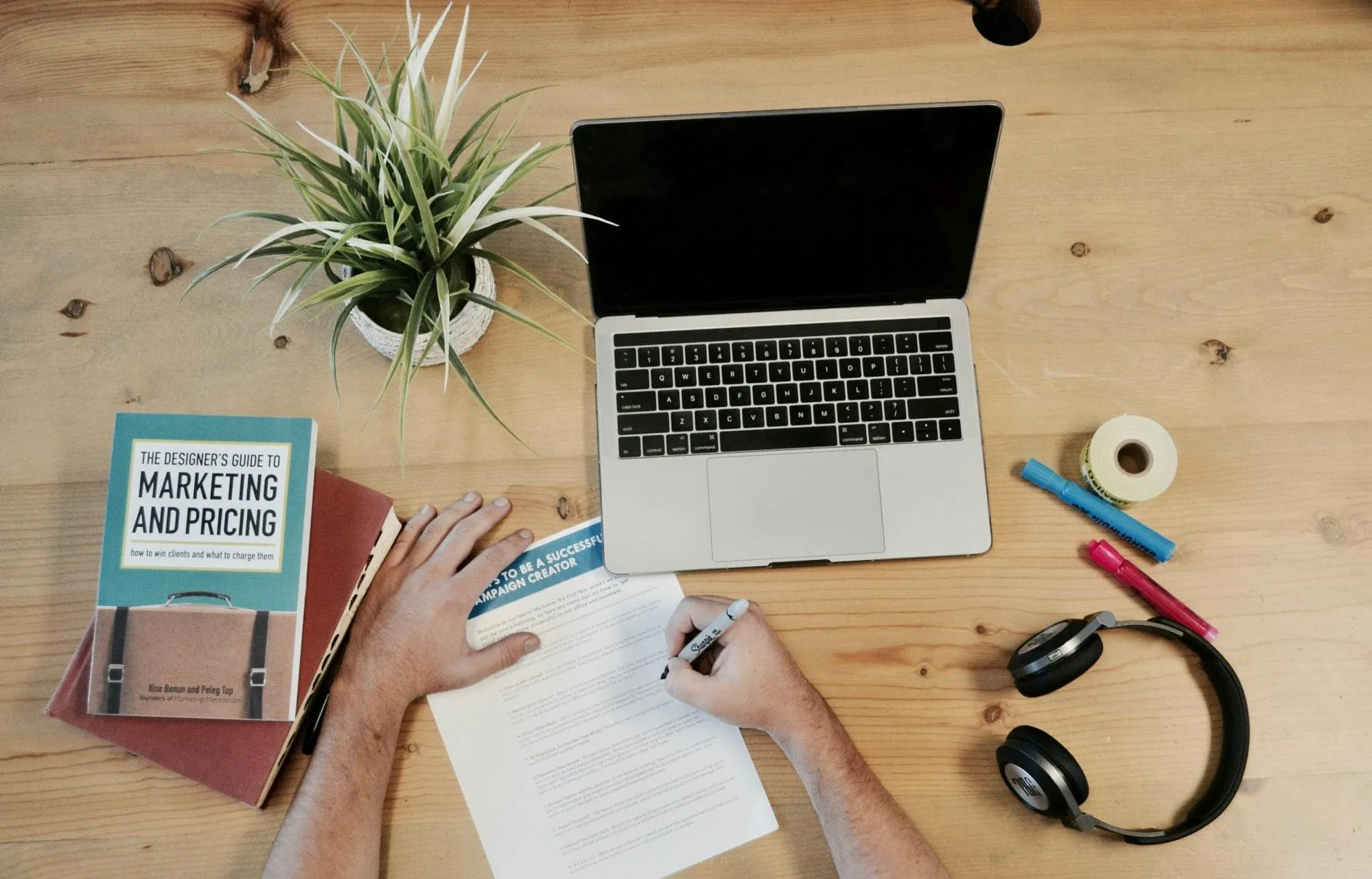 Top view of a workspace with laptop, marketing book, campaign notes, headphones, and writing tools on a wooden desk