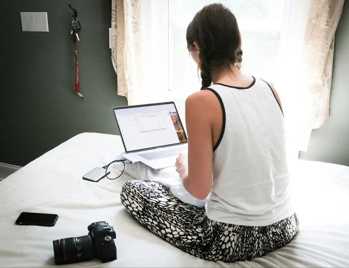 Woman sitting on bed using laptop with DSLR camera and phone nearby, working remotely in natural light