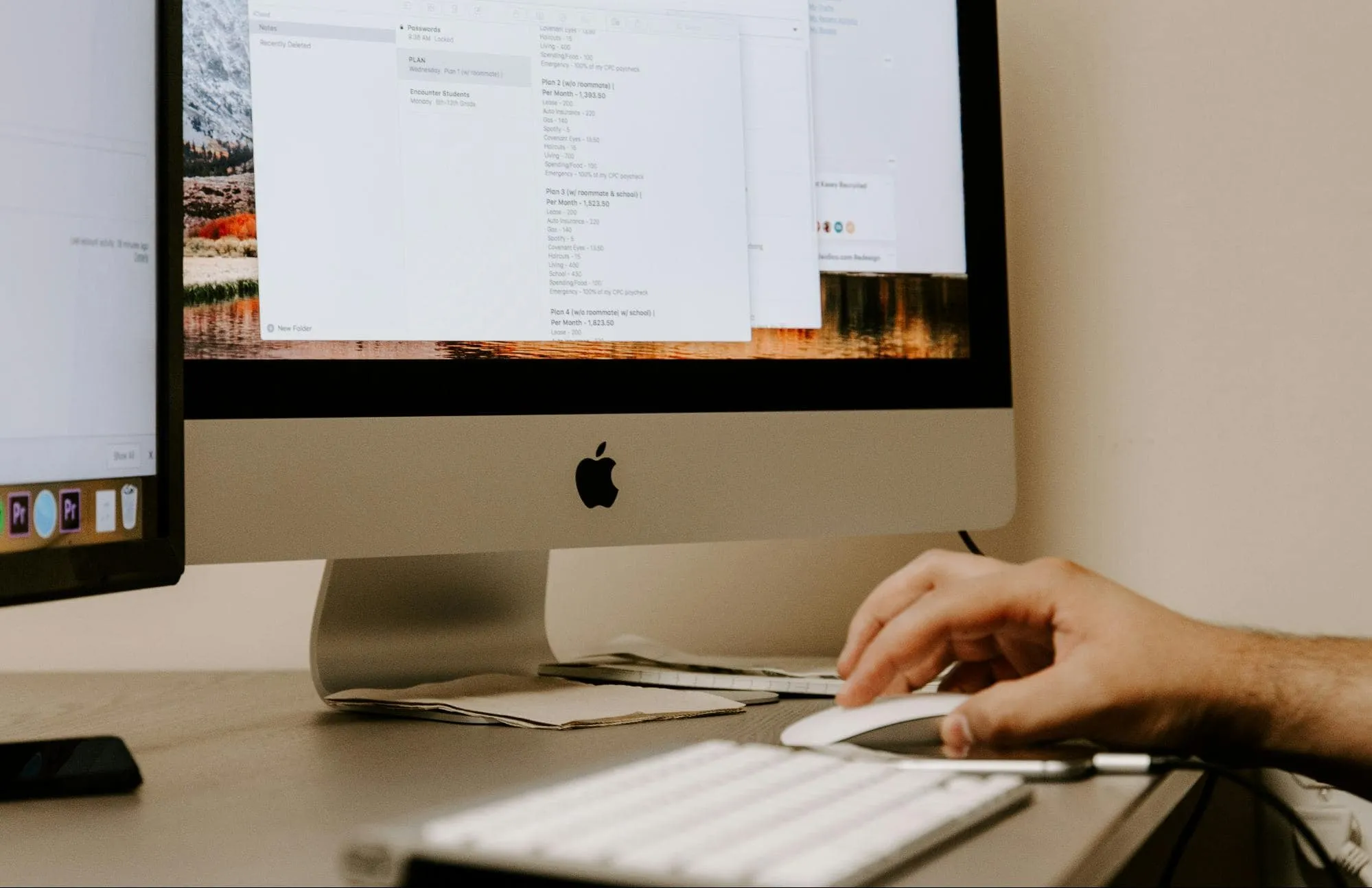 Close-up of a person's hand using an Apple Magic Mouse in front of an iMac screen displaying open documents and desktop folders, part of a dual-monitor workstation setup.