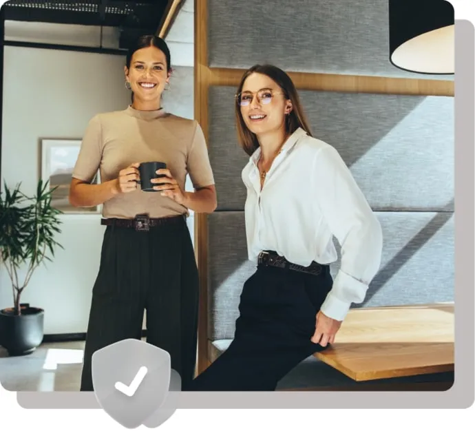 Two confident professional women smiling in a modern office lounge area, one holding a coffee mug and the other leaning casually against a wooden table, symbolizing a positive and secure workplace environment.