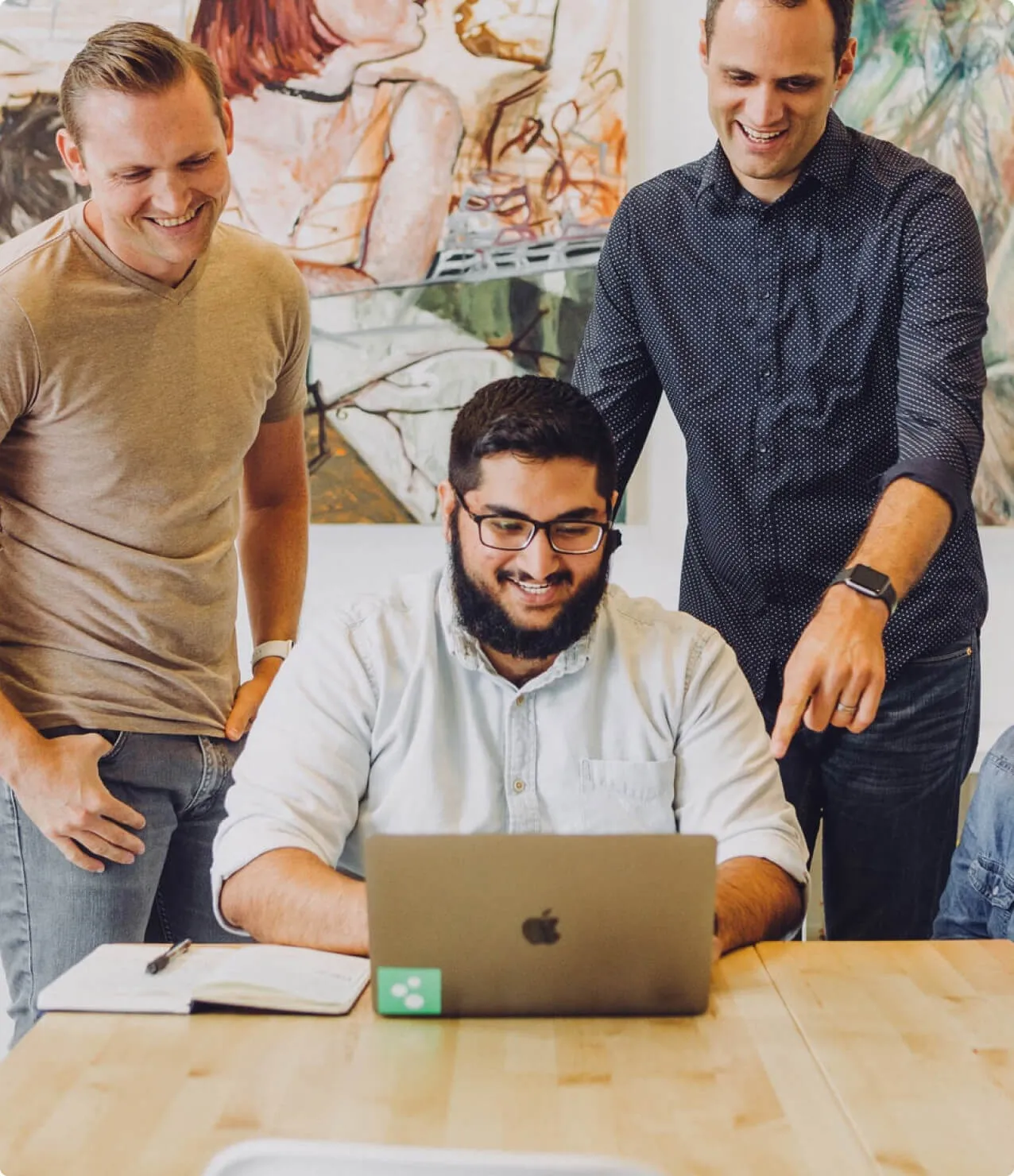 Three men smiling and collaborating around a laptop during a casual office meeting with art on the wall in the background.