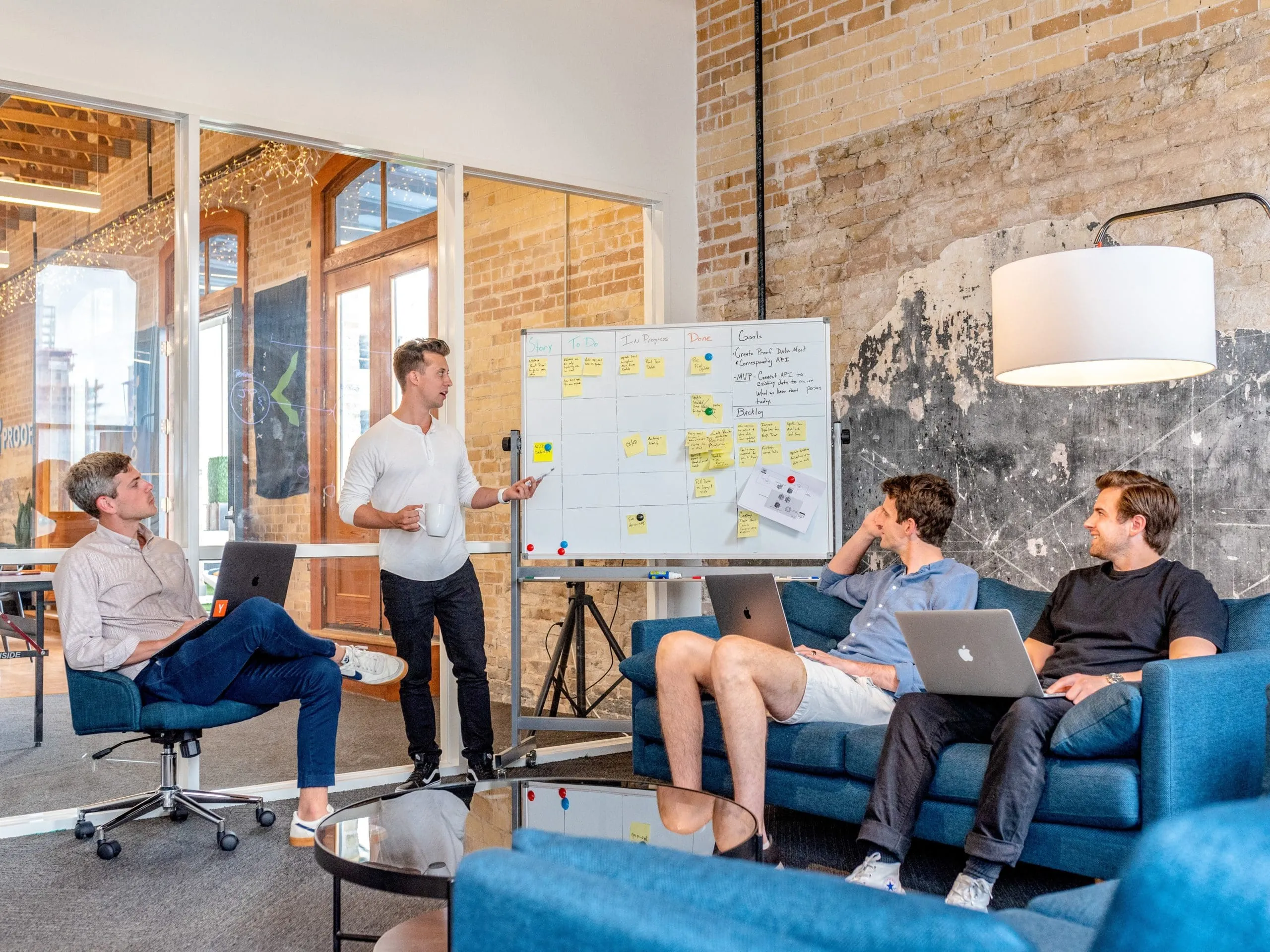 Four men in a modern office space having a team meeting while one of them presents project tasks using a whiteboard covered with sticky notes