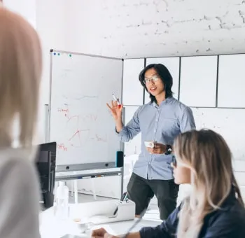 Man presenting marketing data to colleagues at a whiteboard during a team meeting in a bright modern office.