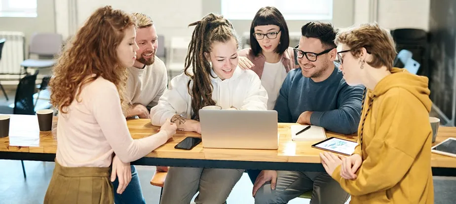 Six people sit around a table, smiling as they review Enterprise SEO Software on a laptop, with coffee cups and notebooks scattered across the table.