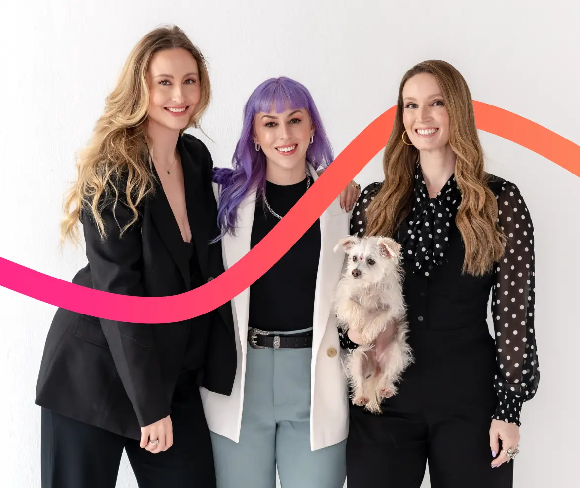 Three women stand together smiling in front of a white background with a curved red and orange line; one woman holds a small white dog.