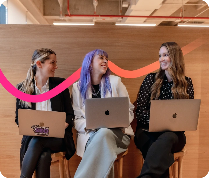 Three women sitting on stools use Apple laptops and smile at each other, brainstorming ideas for enterprise SEO software. A pink wavy line decorates the wood-paneled background.