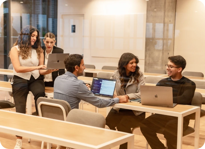Five people in a modern classroom sit and stand around desks, using laptops and discussing Enterprise SEO Software, suggesting a collaborative work or study session.